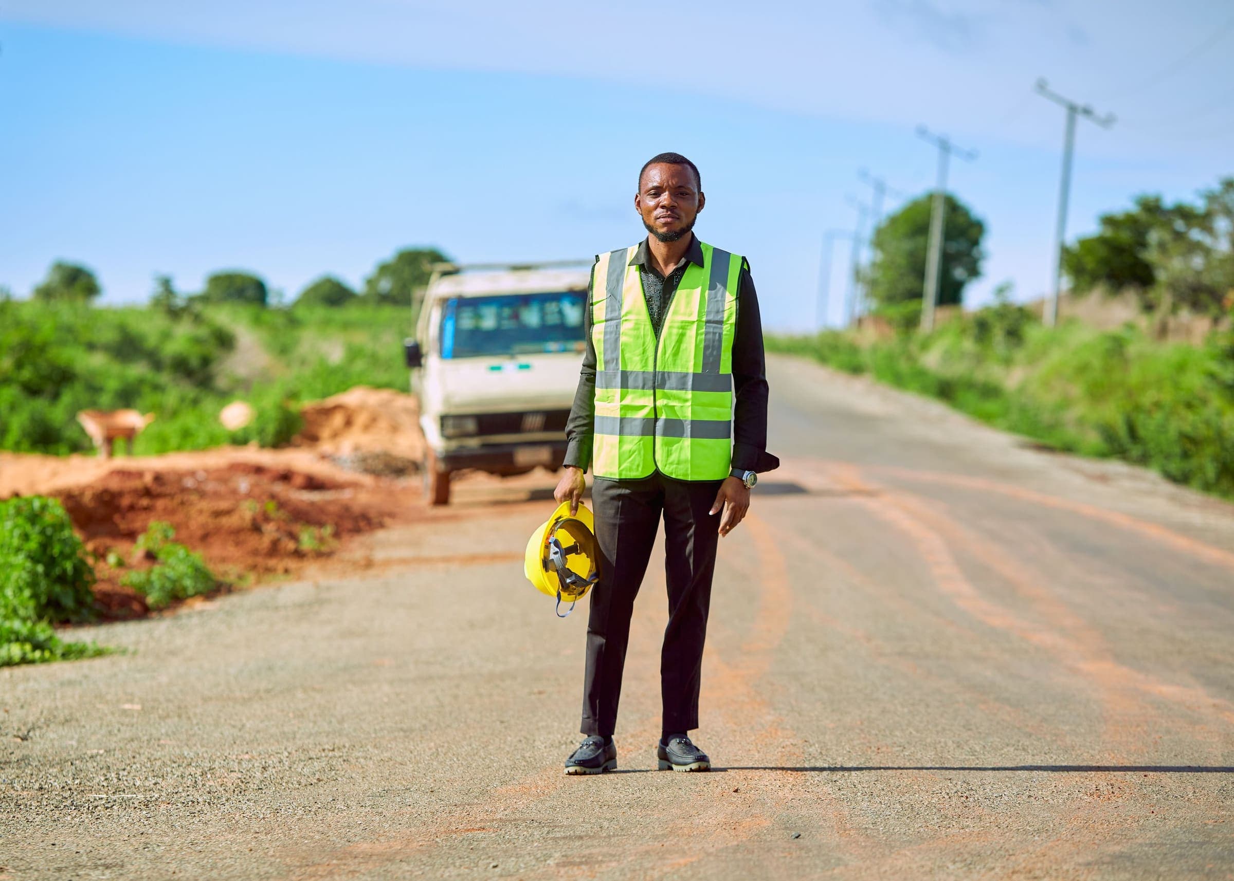 An engineer in a high-visibility vest, holding a hard hat, stands on a rural road beside a parked truck.