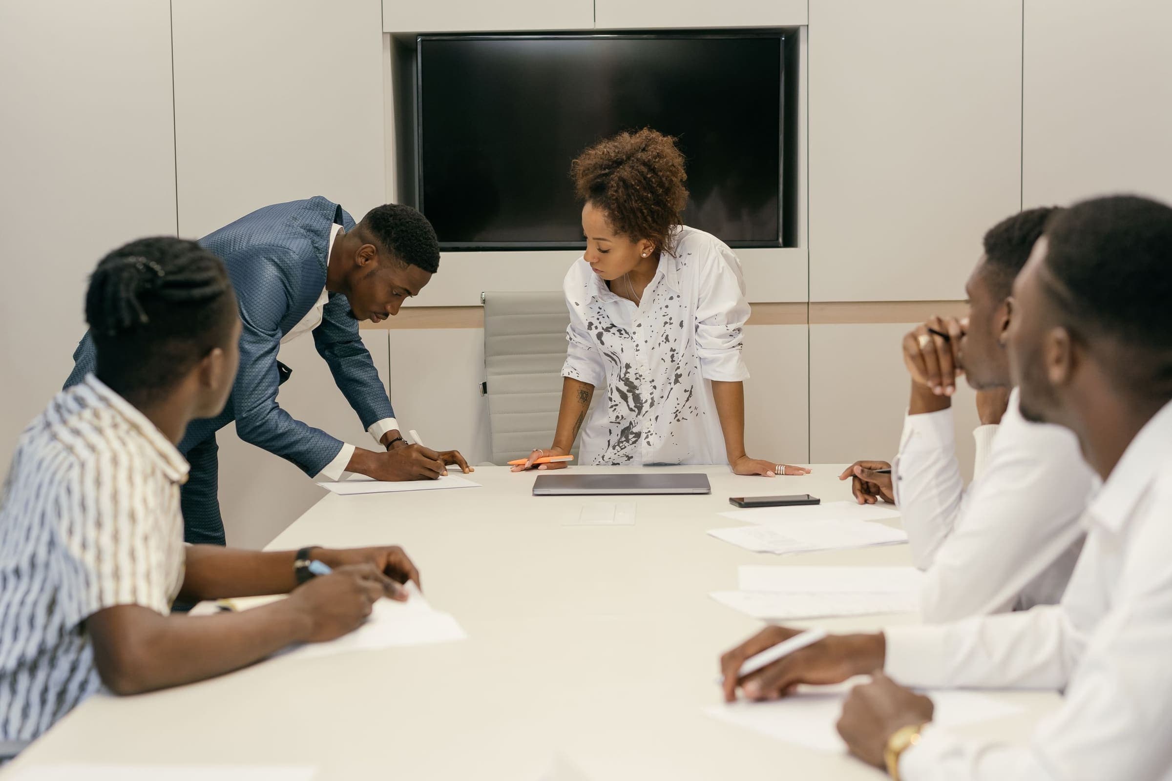 A woman stands at the head of a boardroom table as colleagues review papers and take notes.