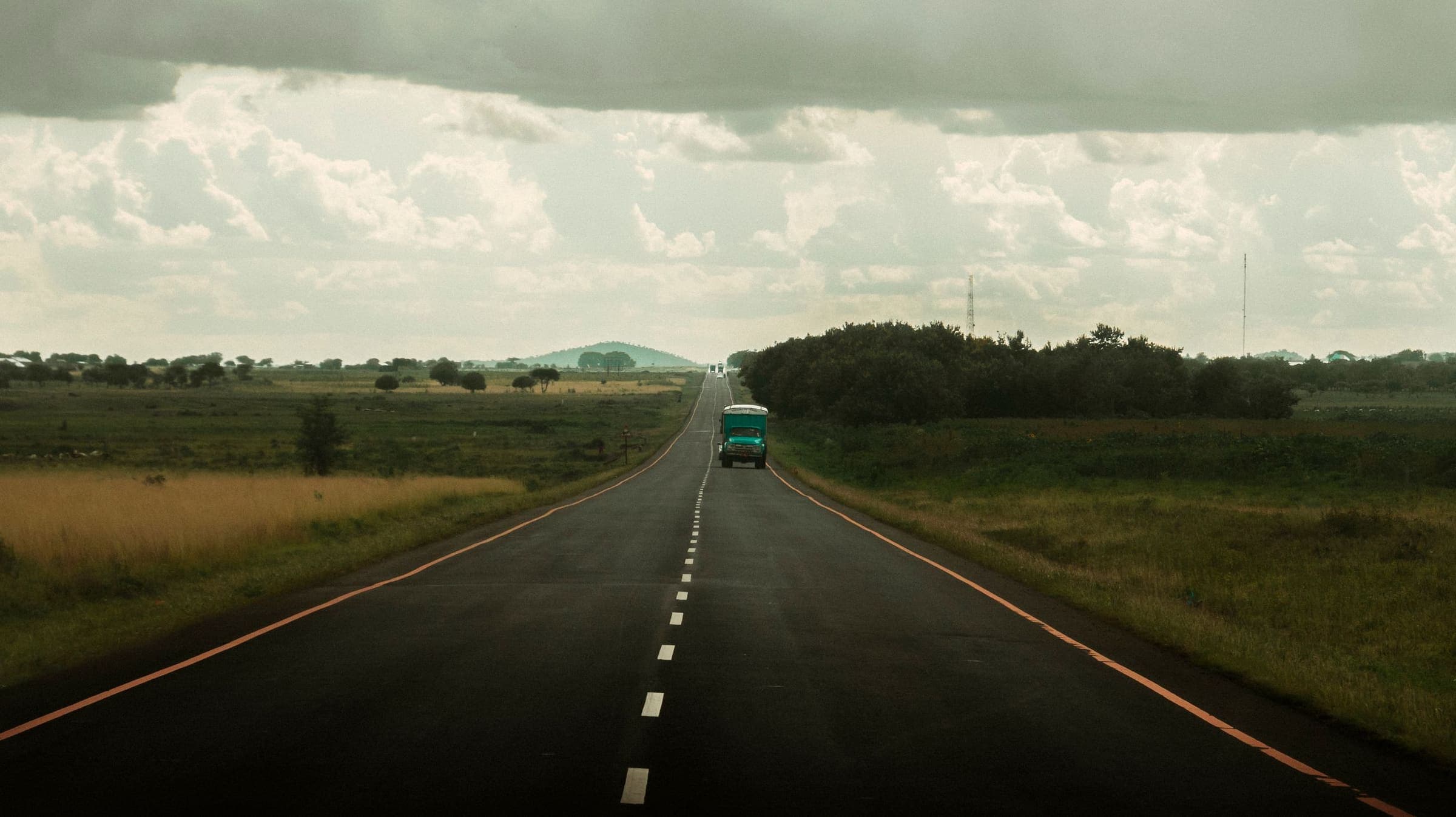 A lone truck on a long, straight highway through open savanna under a heavy sky.