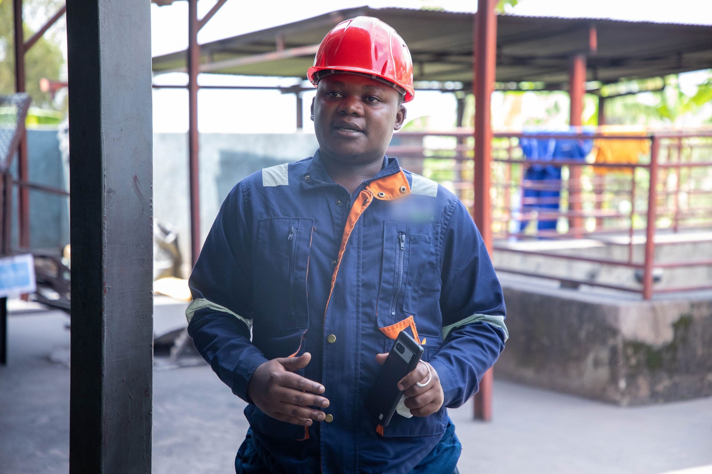 A supervisor in a red hard hat and navy overalls, phone in hand, at a mineral processing plant.
