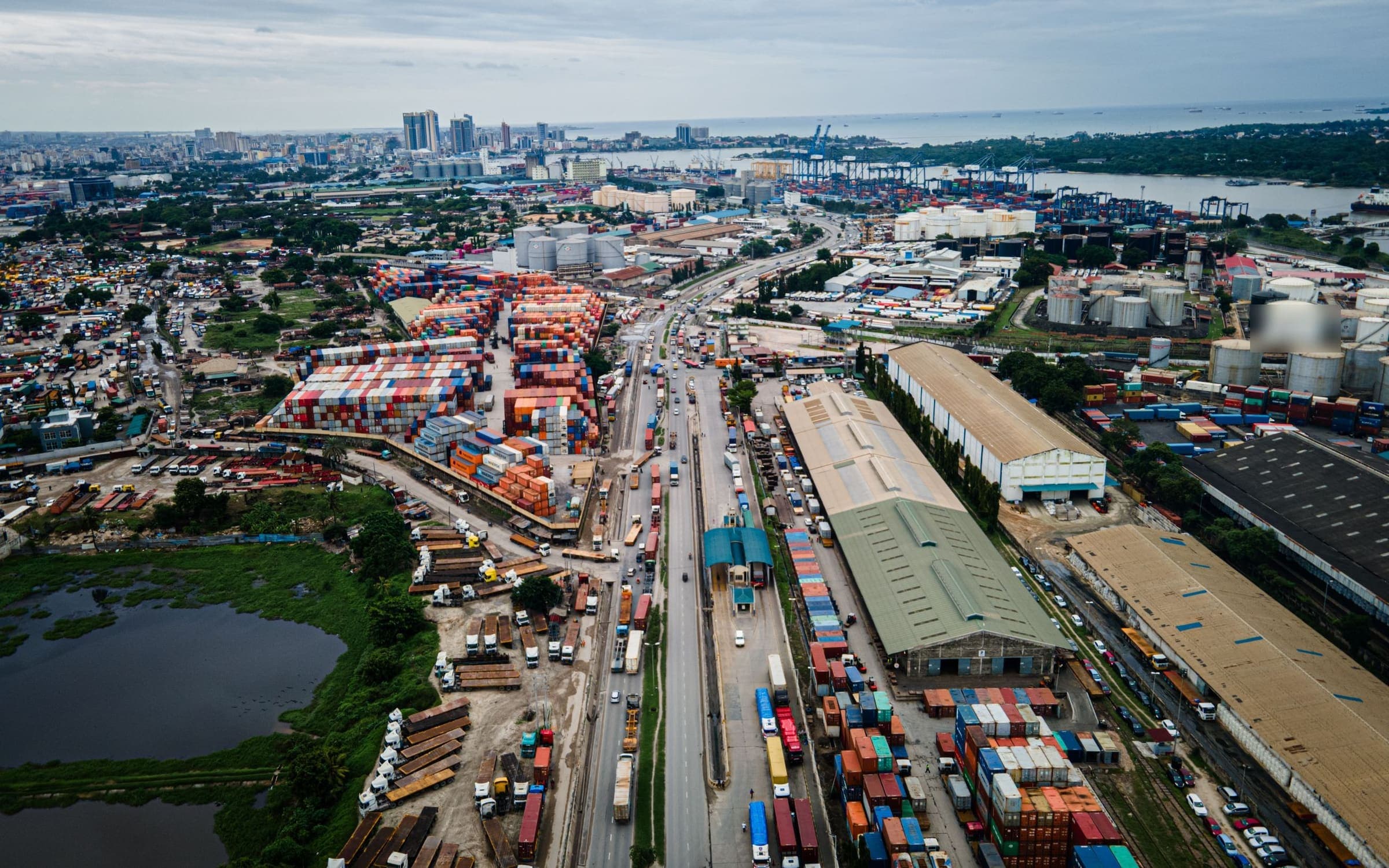 Aerial view of a port-side highway lined with trucks, container depots and warehouses, with harbour cranes beyond.