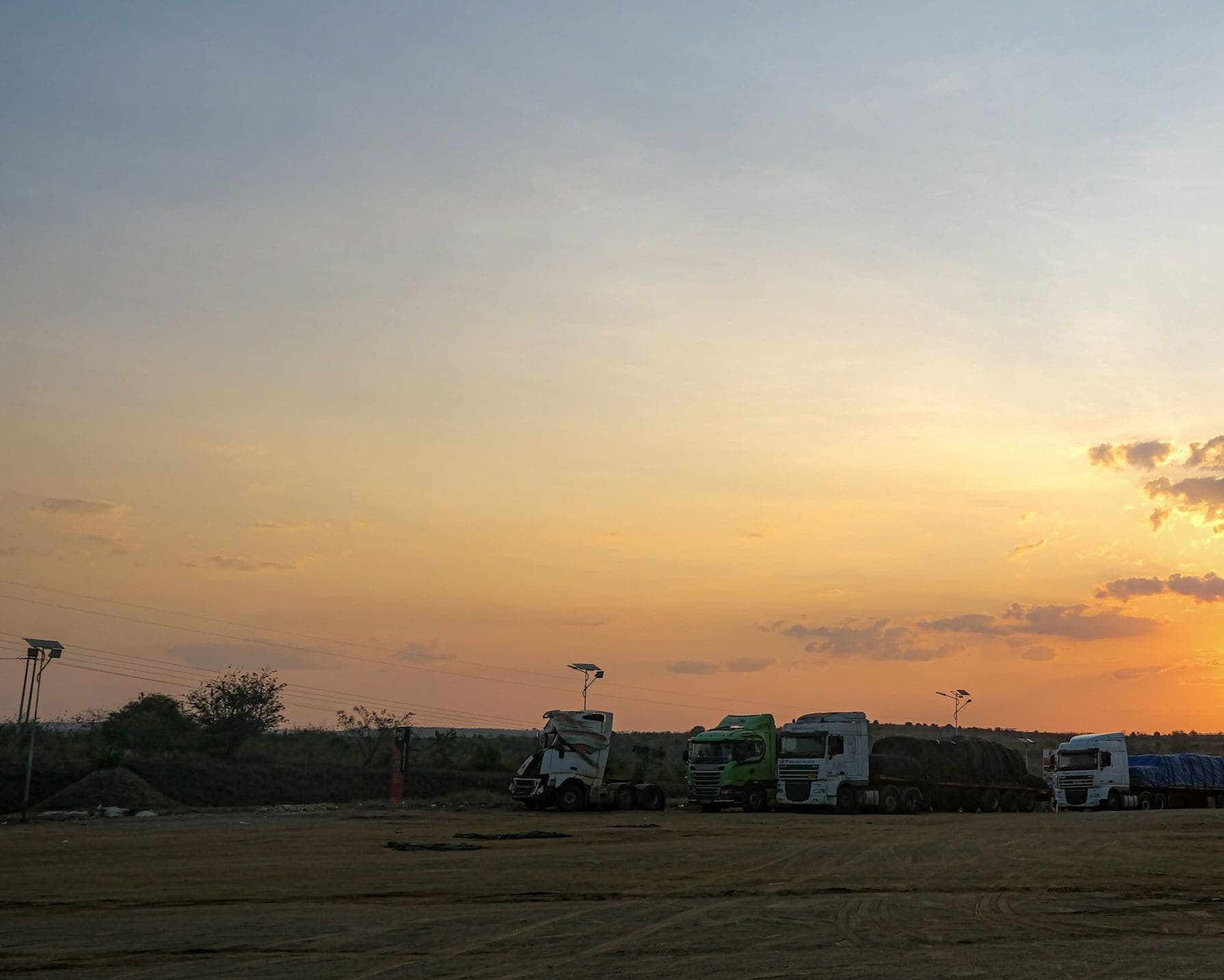 Long-haul trucks parked at a roadside truck stop under an orange sunset sky.